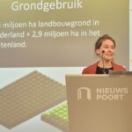 Marga Witteman (witte vrouw, bruin haar, opgestoken in) Staat achter een bokaal waar 'nieuwspoort' op te lezen is. Achter haar is een presentatie slide waar op te lezen is 'GRONDGEBRUIK - 1,3 miljoen ha lanbouwgrond in Nederland + 2,9 miljoen ha in het buitenland'
