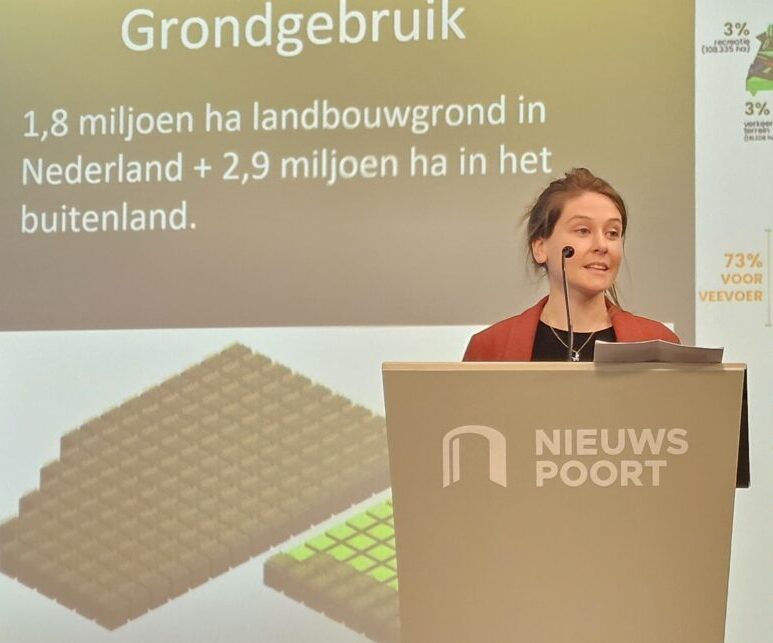 Marga Witteman (witte vrouw, bruin haar, opgestoken in) Staat achter een bokaal waar 'nieuwspoort' op te lezen is. Achter haar is een presentatie slide waar op te lezen is 'GRONDGEBRUIK - 1,3 miljoen ha lanbouwgrond in Nederland + 2,9 miljoen ha in het buitenland'
